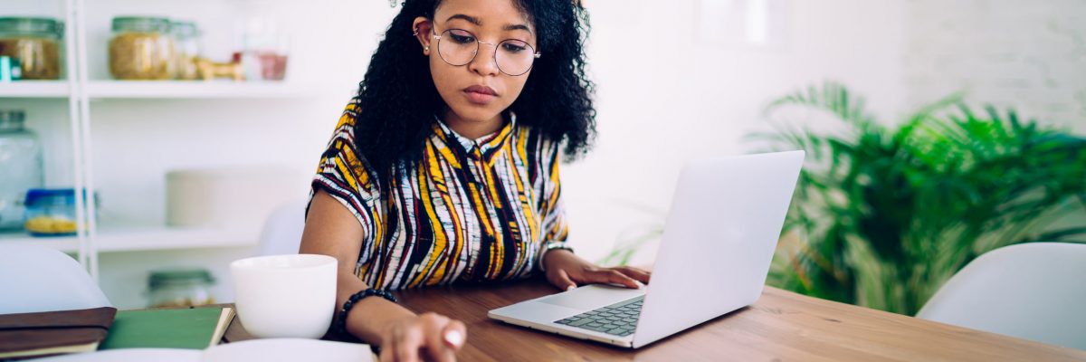 Focused ethnic female freelancer in glasses browsing smartphone and laptop while sitting at table with cup of coffee at home